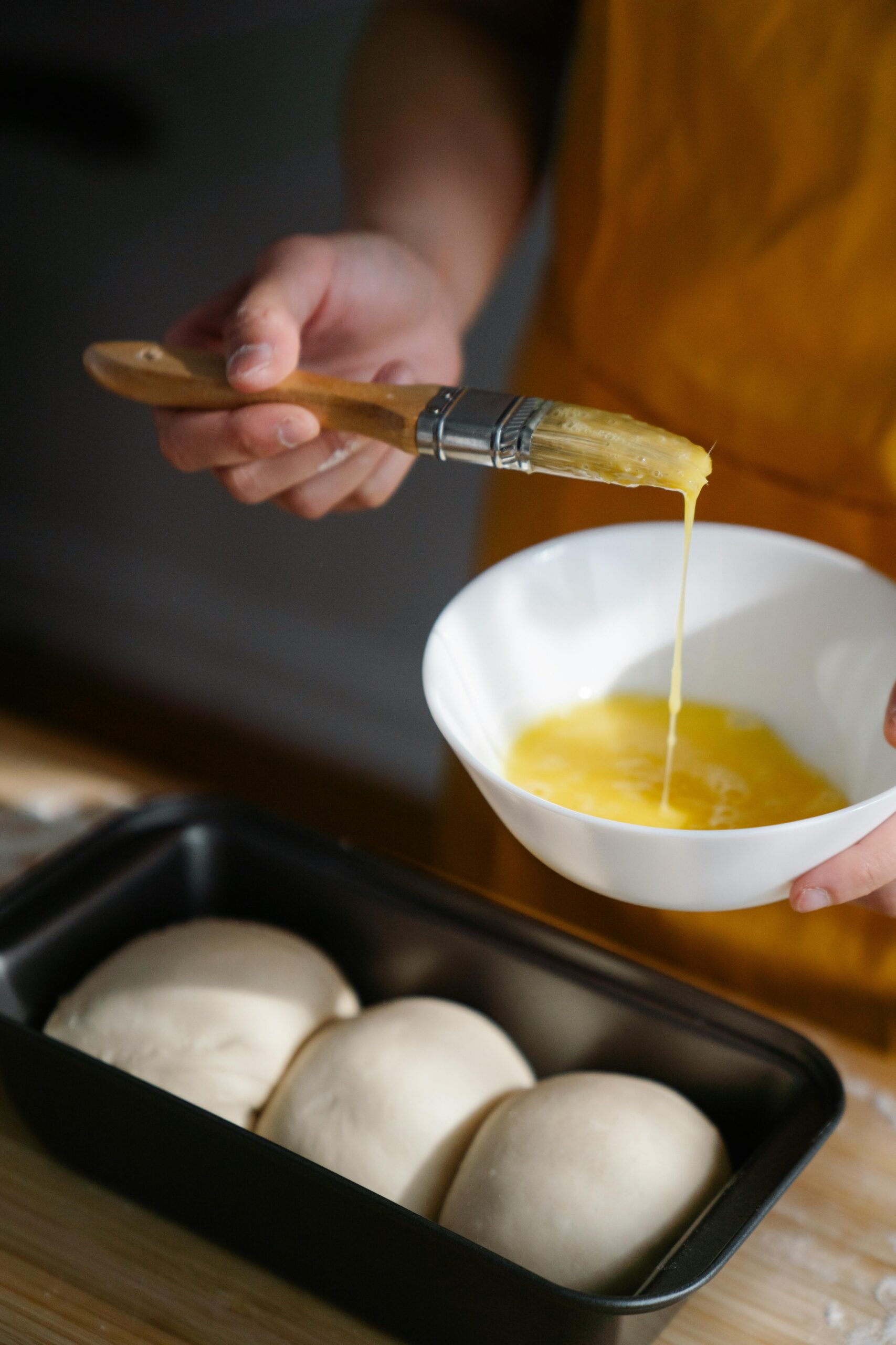 Close-up of hands applying egg wash to dough in a baking pan, creating delicious homemade bread.